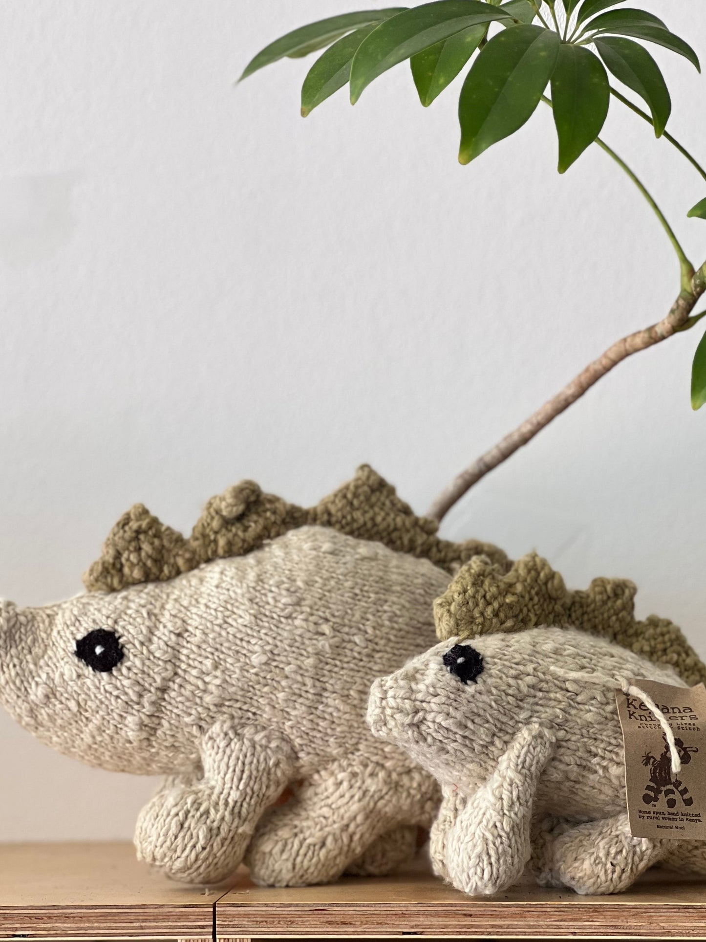 Two knitted dinosaur toys on a wooden shelf with a plant in the background.