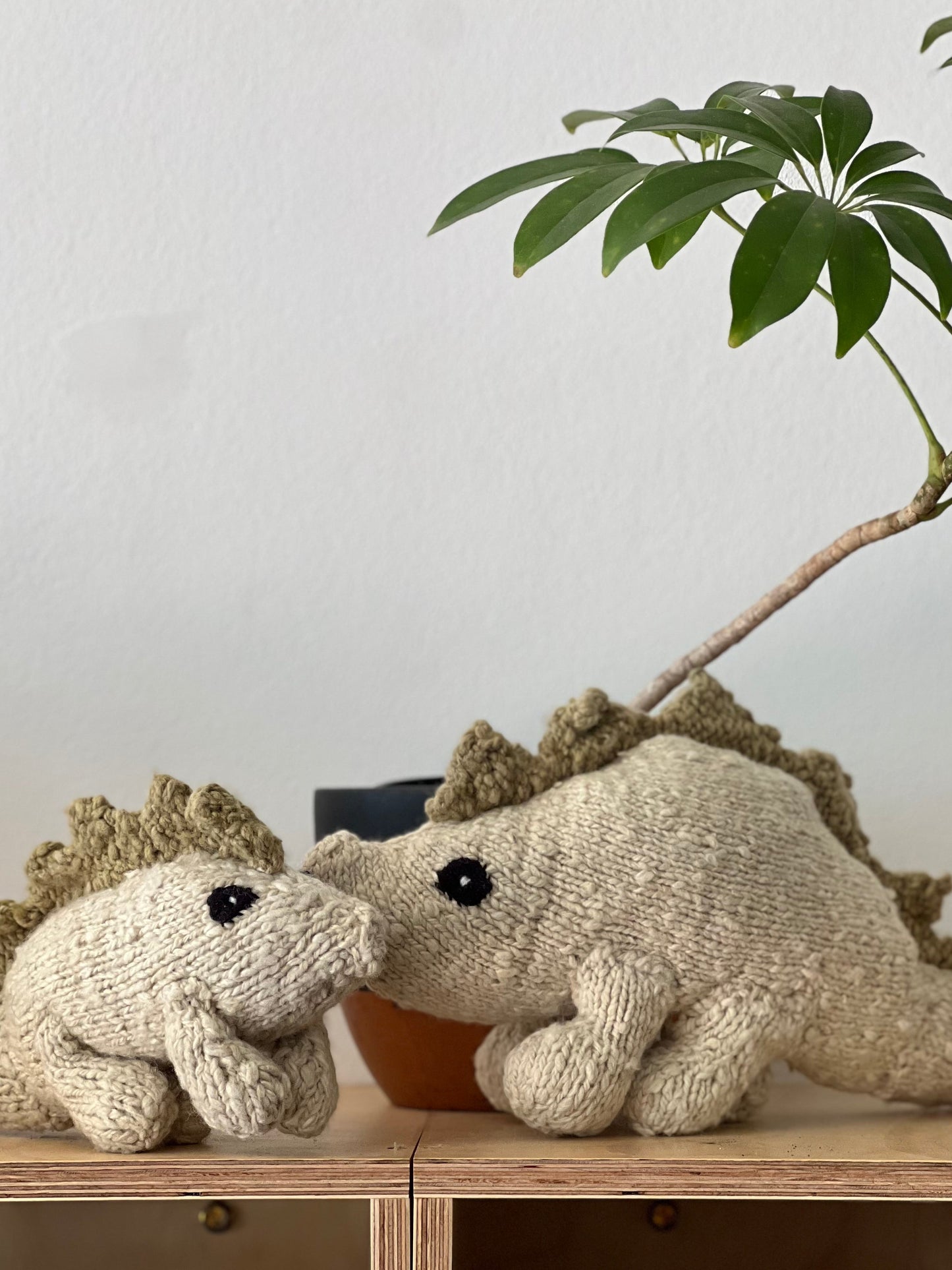 Two knitted dinosaur toys on a wooden shelf with a plant in the background.